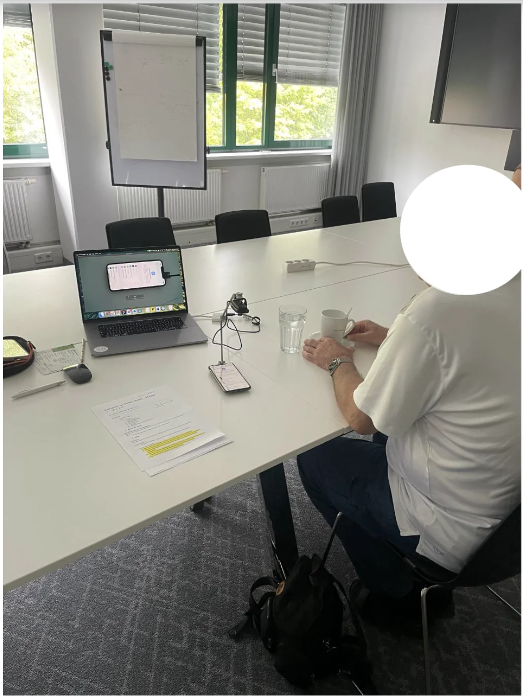 Usability session in Erlangen, August 2025. Participant seated at a conference table with their iPhone on a stand, the app visible on screen. Face anonymised.