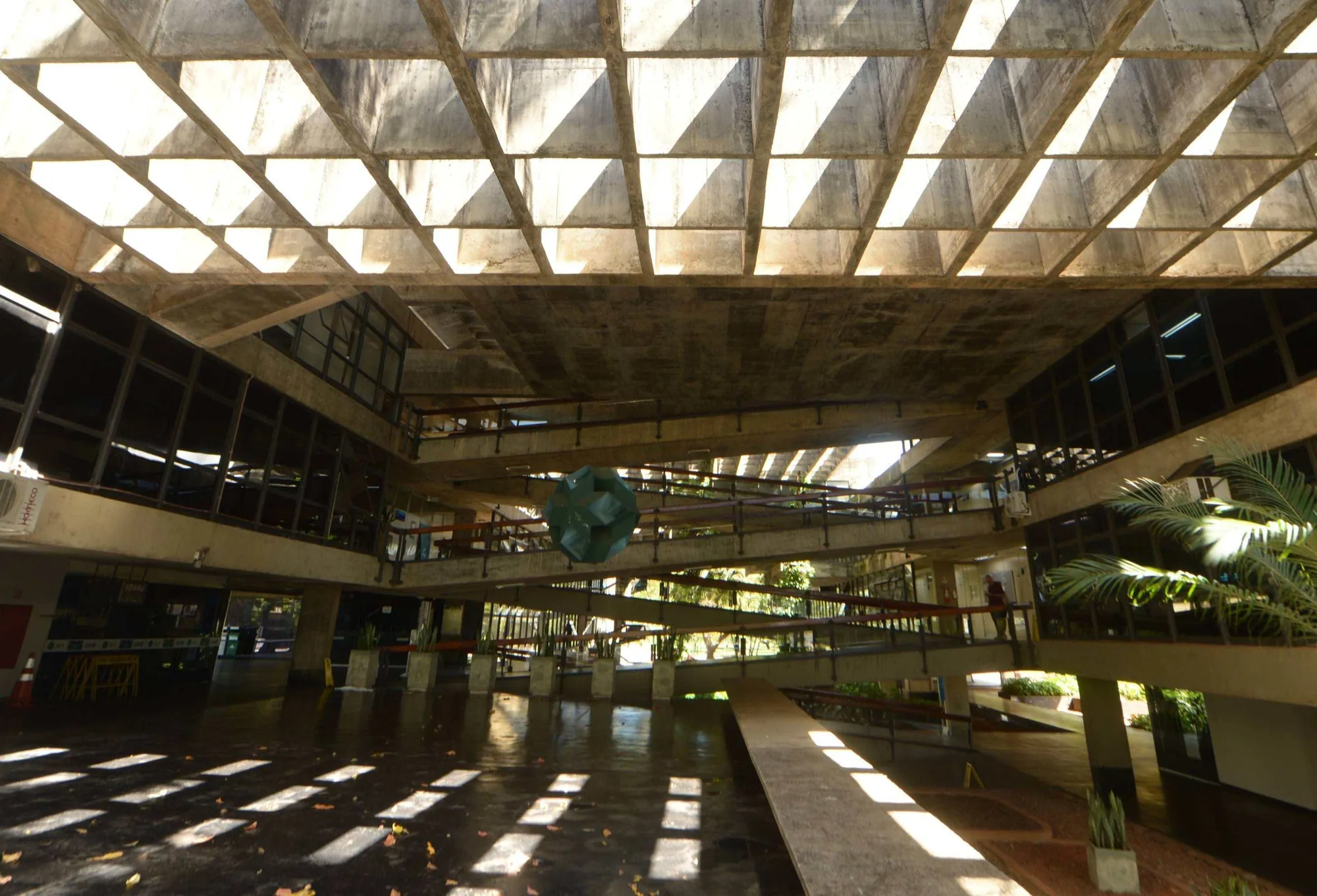 The interior of the Instituto Central de Ciências at the University of Brasília: a vast waffle-slab concrete ceiling on a triangular grid, daylight falling through it onto an open hall, with a green geometric sculpture and palms to one side.